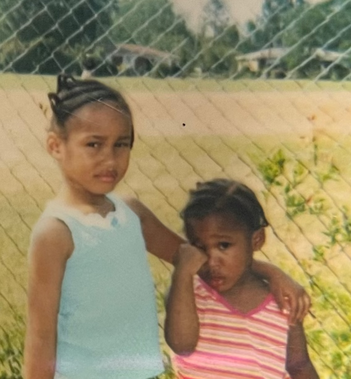 The Hood Enchantress standing with her baby sister in front of a chain linked fence in front of a baseball field. 